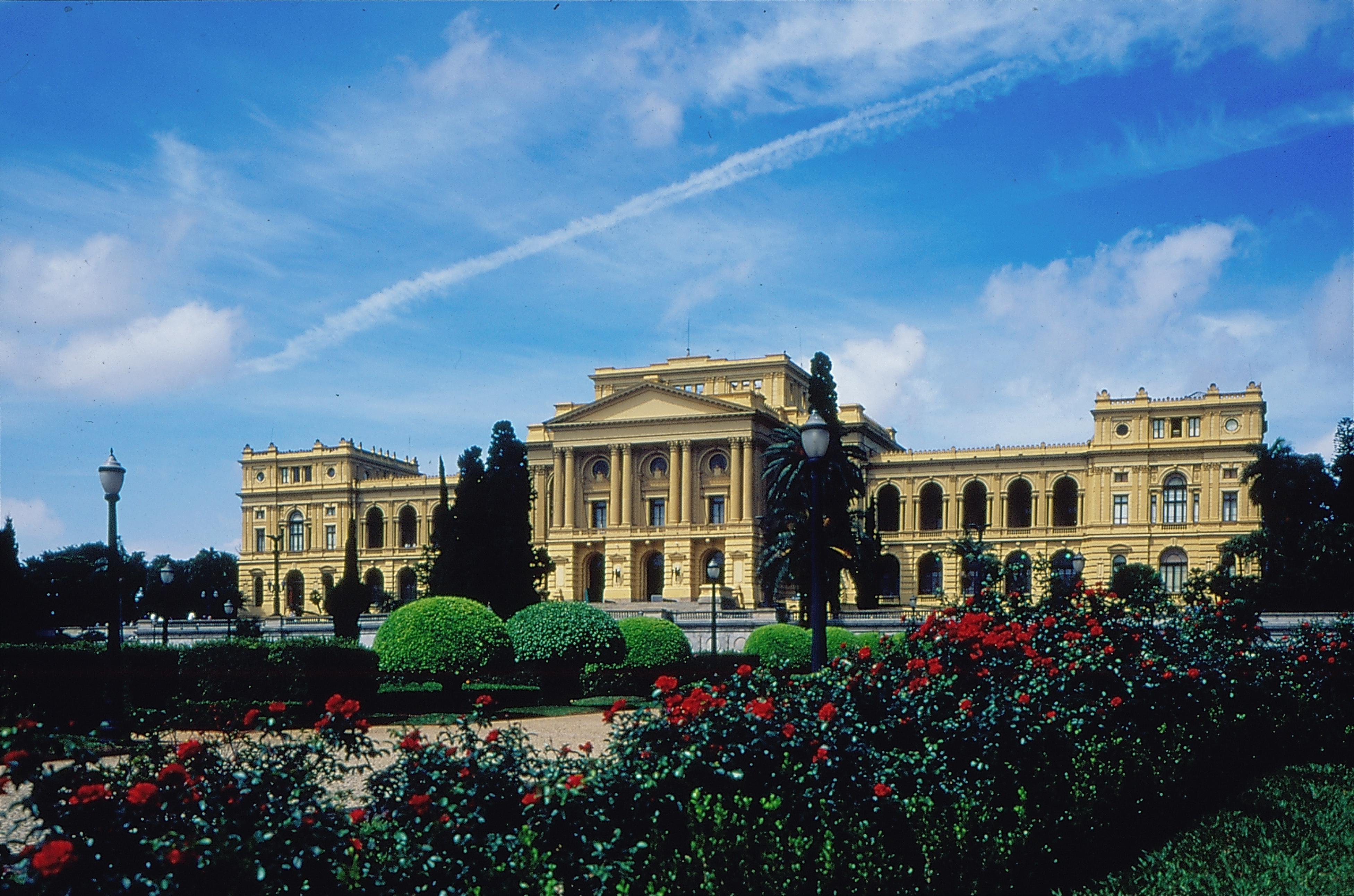 Audiodescrição: Foto da fachada do Museu Paulista. É um imenso edifício de 123 metros de comprimento, 16 metros de profundidade e três andares. O corpo é composto por três torres interligadas por galerias. A frente e as laterais do Museu são compostas por uma imensa área gramada repleta de árvores. No corpo central uma escadaria leva a três portas em arco da entrada principal. Nas laterais, as galerias são fechadas com portas arredondadas de vidro. No primeiro andar, na parte central, há quatro conjuntos de colunas duplas, frisadas e com capitéis decorados de estilo coríntio, que sustentam um frontão triangular com beiral decorado. Os conjuntos de colunas são separados por três portas envidraçadas. No primeiro andar das galerias, as portas são abertas, com arcos, balaústres e colunas decorativas. Acima de cada uma delas há um elemento decorativo formado por uma coroa sobre um escudo e, atrás desse elemento, um óculo: abertura redonda com vidro. À frente do Museu há um imenso jardim que mistura um gramado, diferentes tipos de árvore, flores vermelhas e alguns postes de luz. Ao fundo há um céu de um azul intenso, repleto de nuvens brancas.